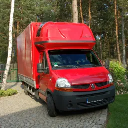 Czerwony samochód dostawczy Renault z plandeką, zaparkowany na brukowanej posesji przed domem, z widocznym napisem 'MOBILE HOME' na nadbudowie kabiny.